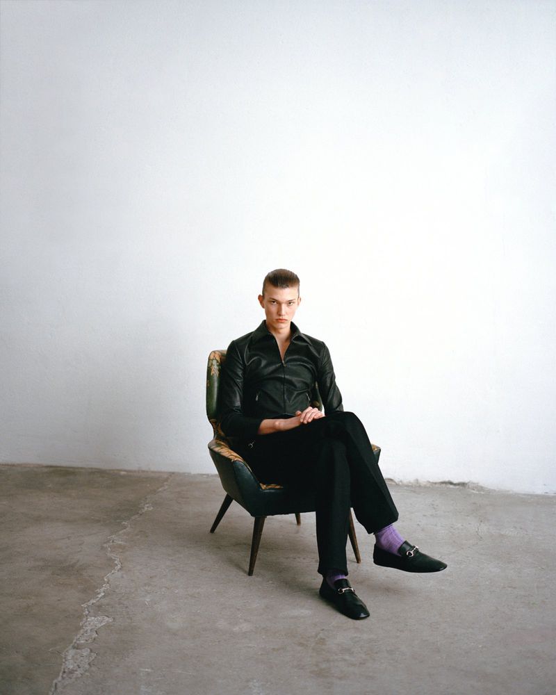 A young adult male in a black leather jacket and black trousers sits in a vintage armchair against a minimalist white backdrop in a studio.