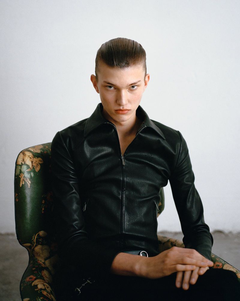 Portrait of a young adult male with slicked-back hair, wearing a black collared zip-up leather jacket, and seated in a floral armchair.