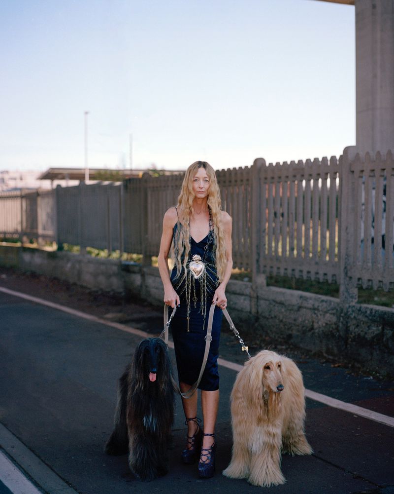 Woman walks two Afghan Hounds on a road near concrete structures, wearing a dark dress, strappy heels, and has long blonde hair.