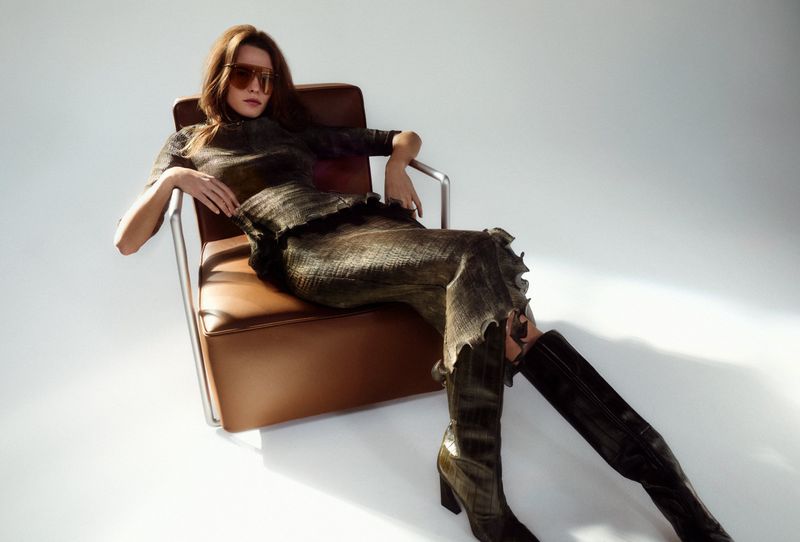 A young adult female model in a brown, patterned dress and black boots is sitting in a leather chair against a white backdrop.