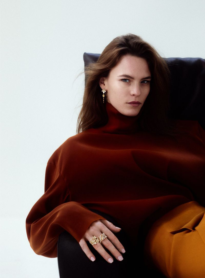 Brunette woman poses in chair wearing a rust-colored turtleneck, gold jewelry, and orange pants against a white backdrop.
