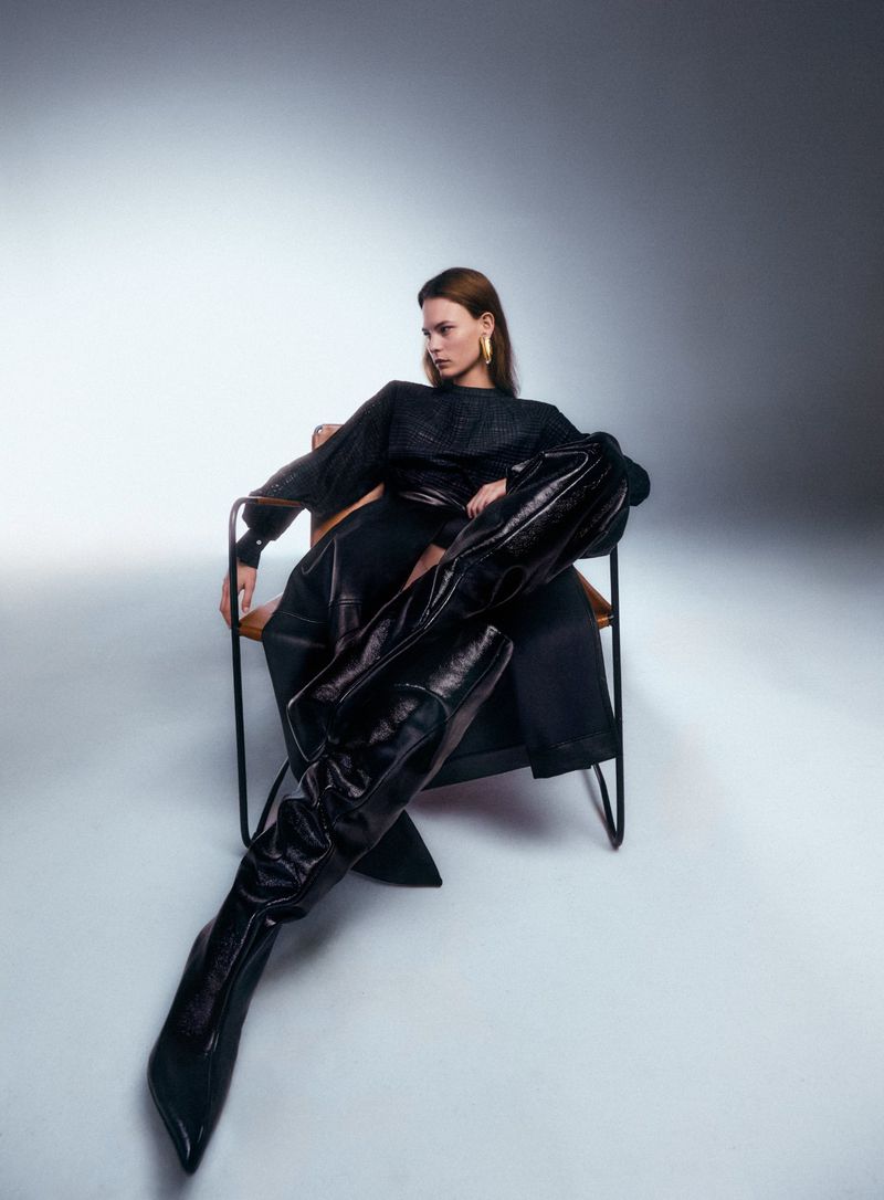 Model Coline Leclere seated in a modern chair wearing exaggerated black thigh-high boots and a black outfit against a studio backdrop.