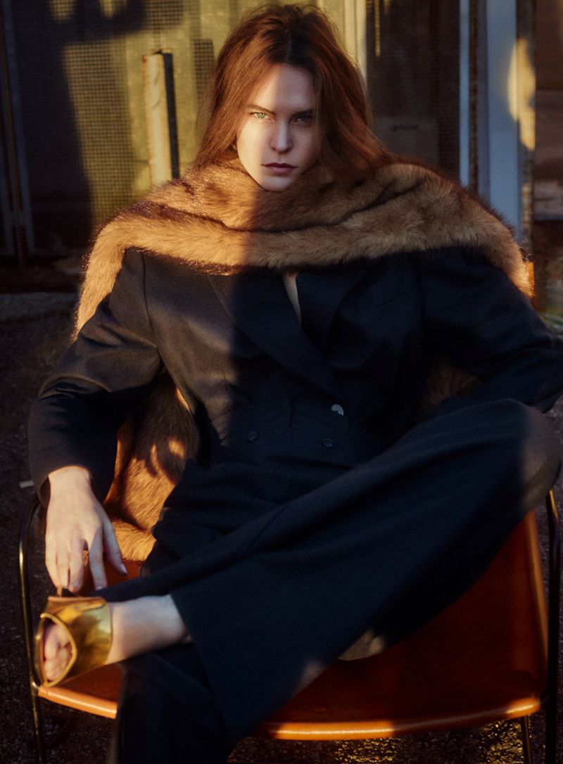 Fashion portrait of a woman with long auburn hair wearing a black suit and brown shawl, seated outdoors in golden hour lighting.