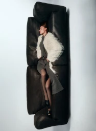 Model lying on a black leather sofa wearing a grey dress and white fluffy coat against a white backdrop.