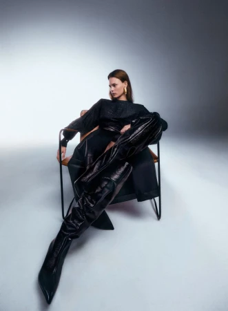 Model Coline Leclere seated in a modern chair wearing exaggerated black thigh-high boots and a black outfit against a studio backdrop.