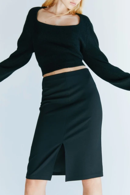 Woman in black sweater and skirt poses against a white backdrop in a fashion shot, with a medium shot capturing her upper body and legs.