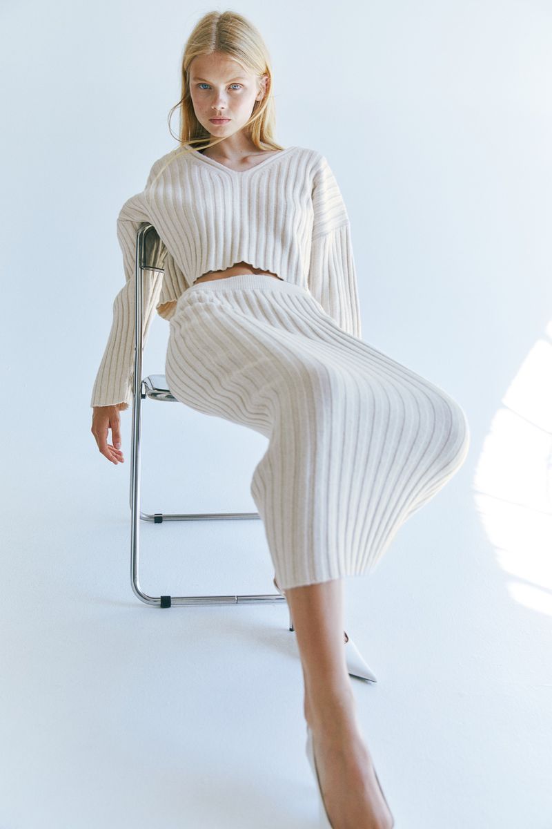 Young adult woman posing indoors, seated on a metal chair in a knitted cream-colored sweater and skirt; fashion-focused shot.