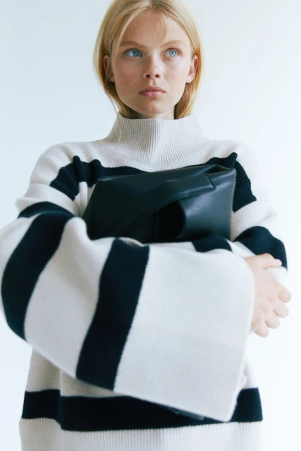 A blonde woman wearing a black and white striped turtleneck sweater holds a black clutch in a studio setting with soft, diffused lighting.
