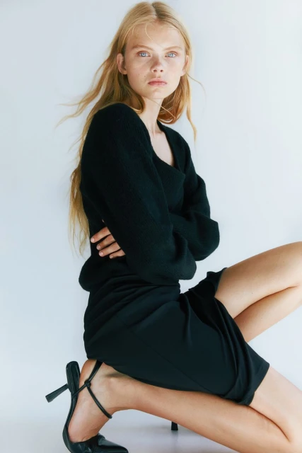 A blonde fashion model poses kneeling in a black long-sleeve top, black skirt, and black high-heels against a white background in a photo studio.