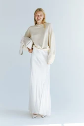 Model in a cream knit sweater and white satin skirt holding a light handbag against a plain background
