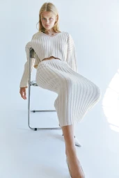 Model in ribbed cream knit sweater and matching skirt seated on metal chair against white seamless background