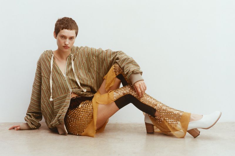 Young woman with short hair poses in a studio wearing a striped hoodie, embellished skirt, leggings, and heels against a white background.