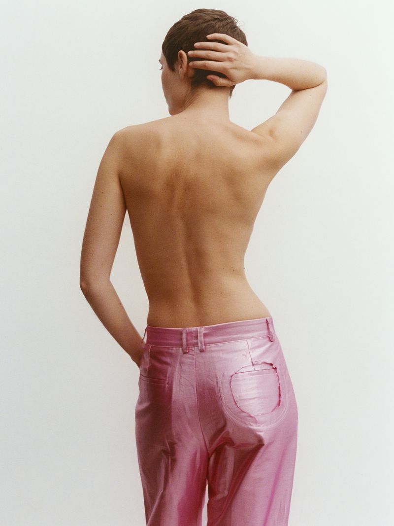 Back view of a woman with short hair posing with a hand on her head, wearing pink pants in a light studio setting.