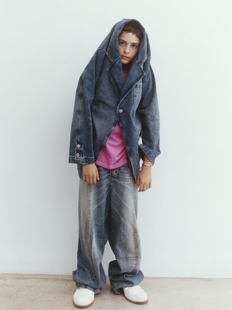 Young person wearing oversized denim jacket and baggy jeans, standing against a white wall in an indoor studio setting.