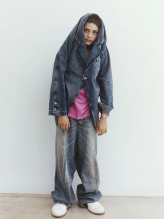 Young person wearing oversized denim jacket and baggy jeans, standing against a white wall in an indoor studio setting.