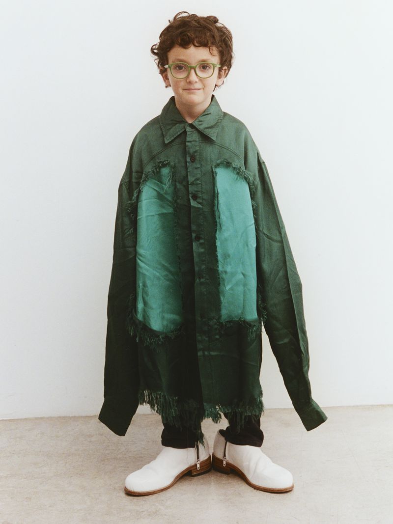 A child with curly hair, wearing glasses and an oversized green shirt with white boots, stands in front of a plain white wall.