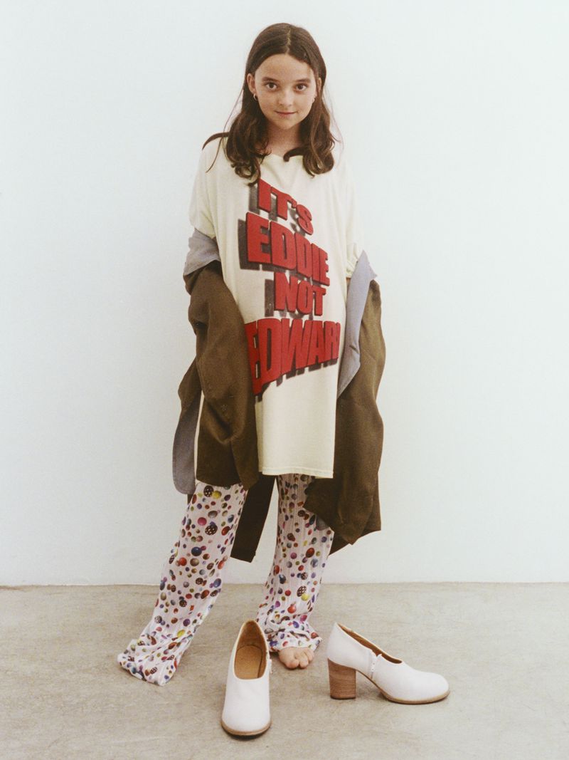 Young model in oversized t-shirt and patterned leggings poses with stylish white pumps against a neutral background, fashion-forward.