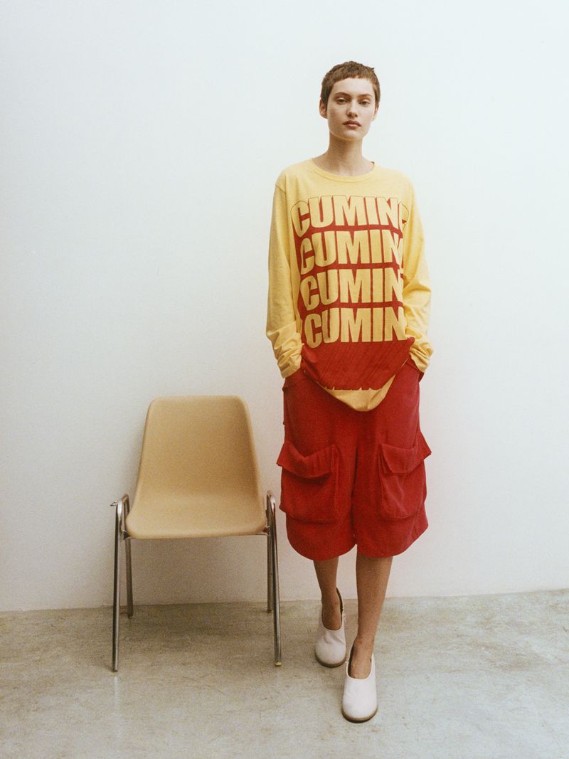 A person with short hair is walking in a yellow long sleeve shirt and red shorts while standing next to a beige chair.