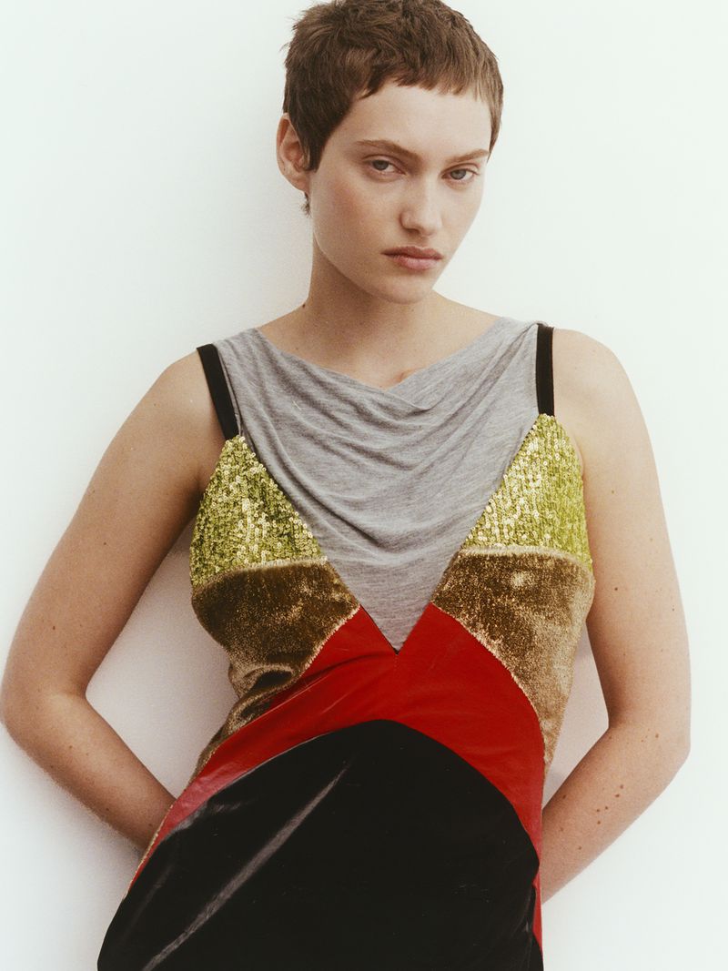 Fashion portrait of a woman with short brown hair wearing a colorful patchwork dress with gold, brown, red, and black tones in a studio setting.