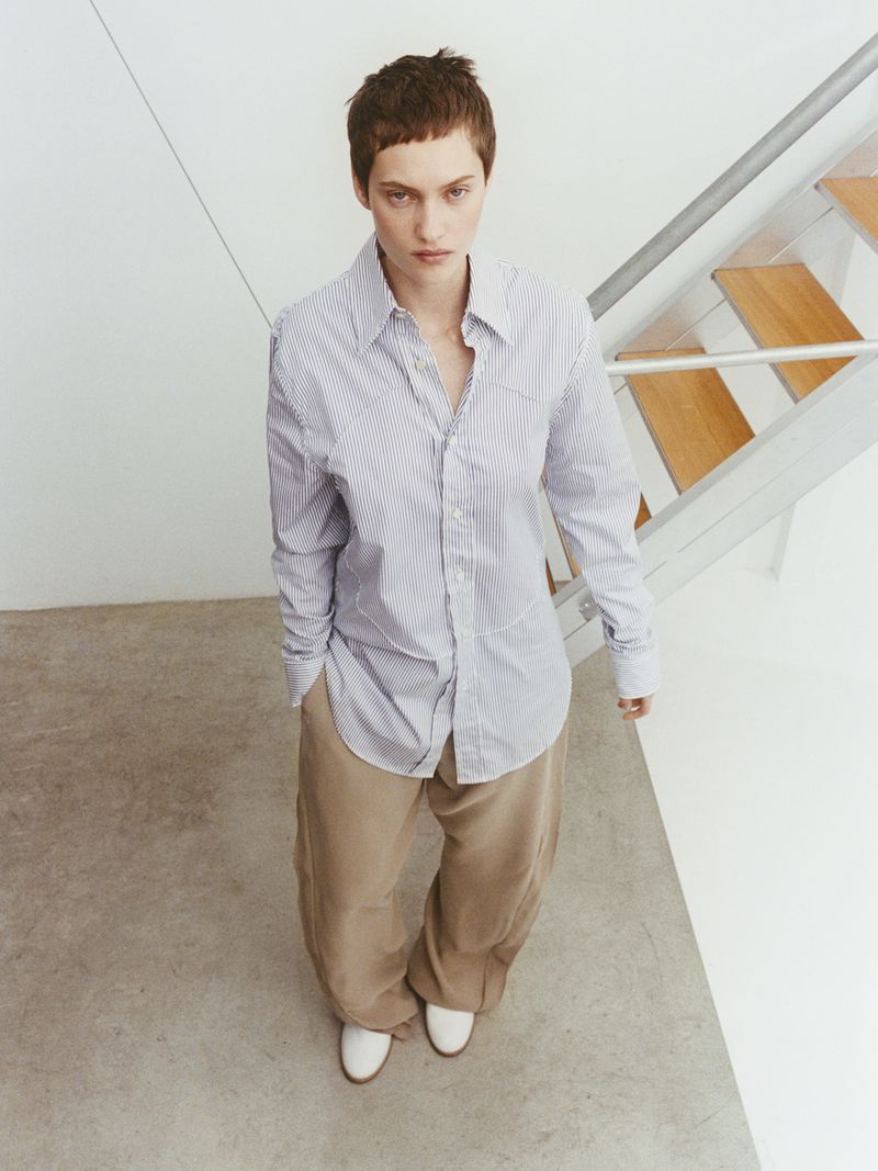 Overhead shot of a young adult woman in a striped shirt and loose pants, standing in an indoor space with staircase in background.