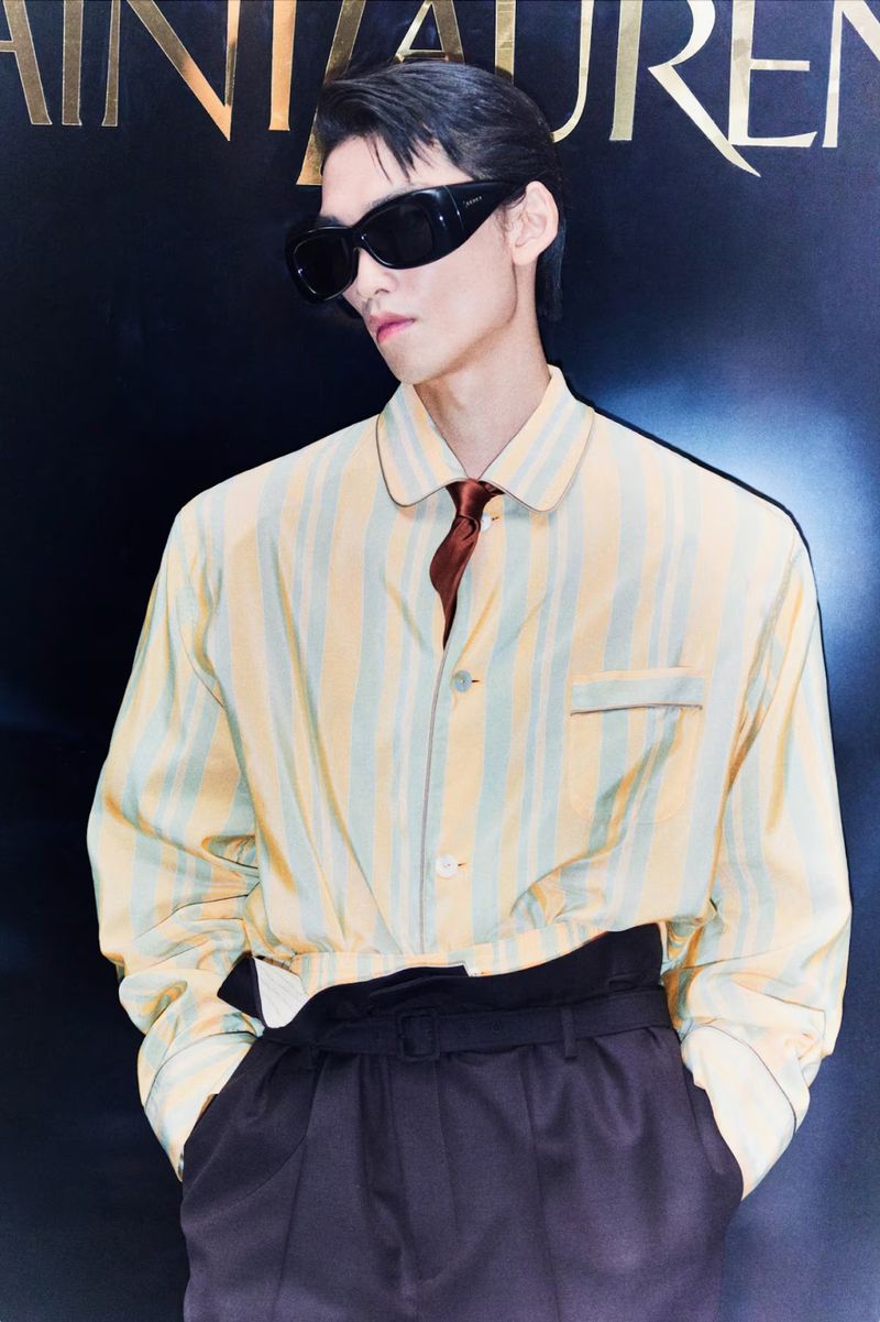 Portrait of an adult male wearing sunglasses, a striped shirt, and dark pants in front of a 'Saint Laurent' branded background.