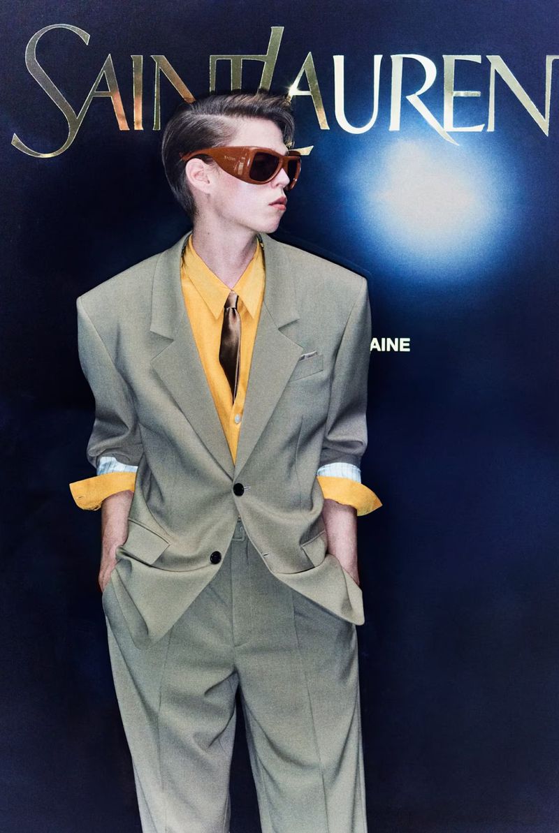 Stylish adult poses in a sage suit, accessorized with a yellow shirt and retro sunglasses, against a high-fashion branded backdrop.