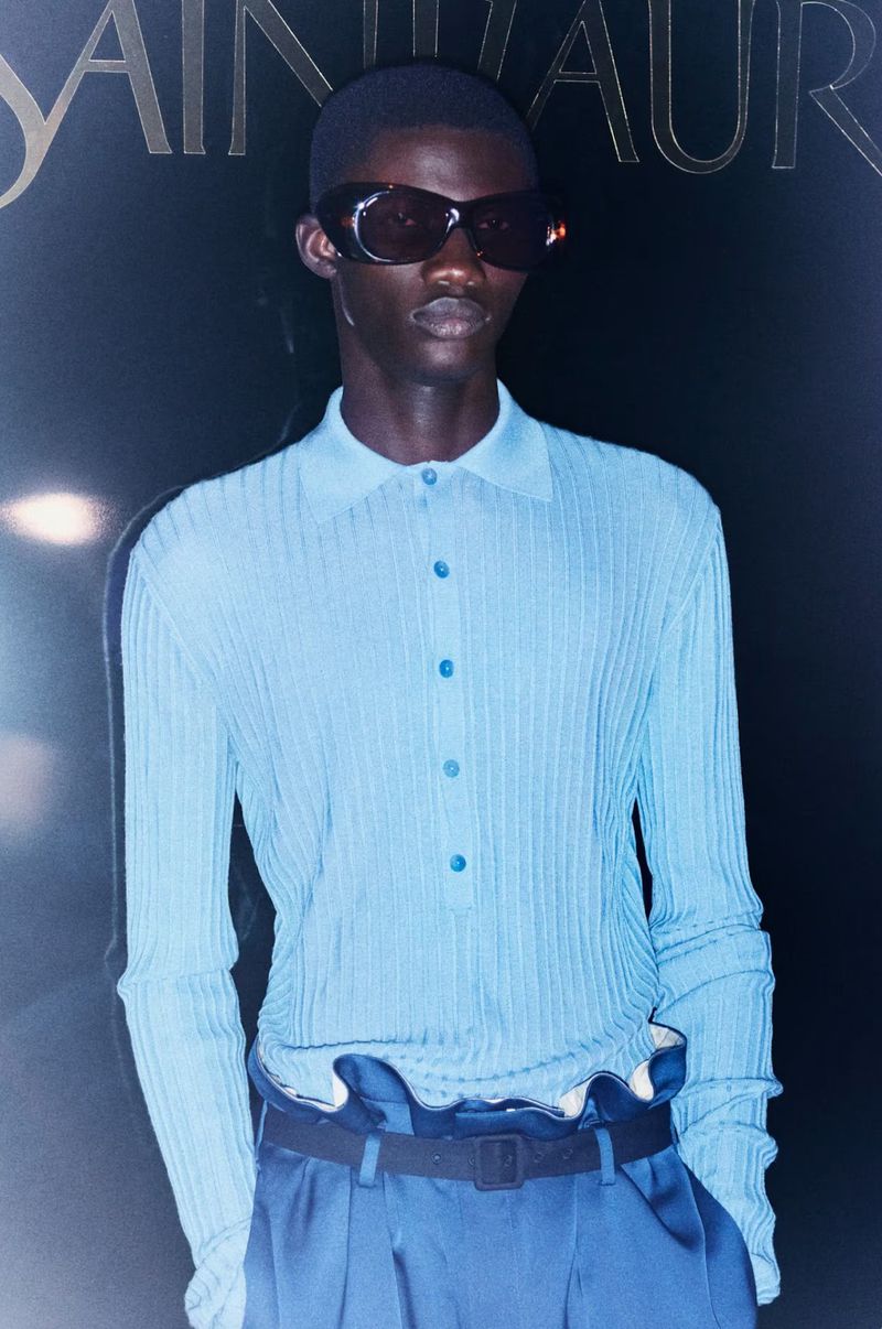 A dark-skinned male model in a light blue button-down knit shirt, sunglasses, and blue pants poses in front of a logo backdrop.