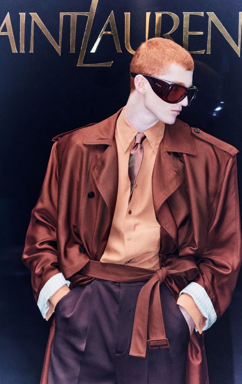 Fashionable male model posing in a brown belted trench coat and sunglasses in front of a Saint Laurent logo, creating a stylish aesthetic.