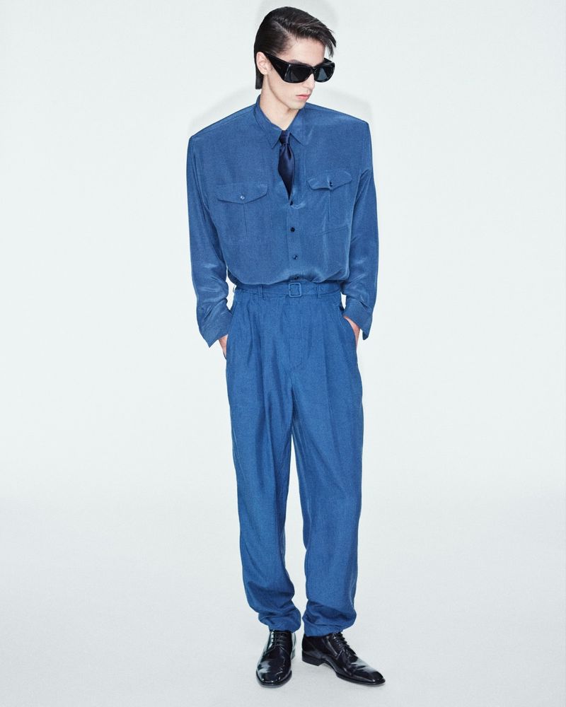 Stylish adult male posing in matching blue shirt and trousers against a white backdrop, accessorized with sunglasses and black leather shoes.