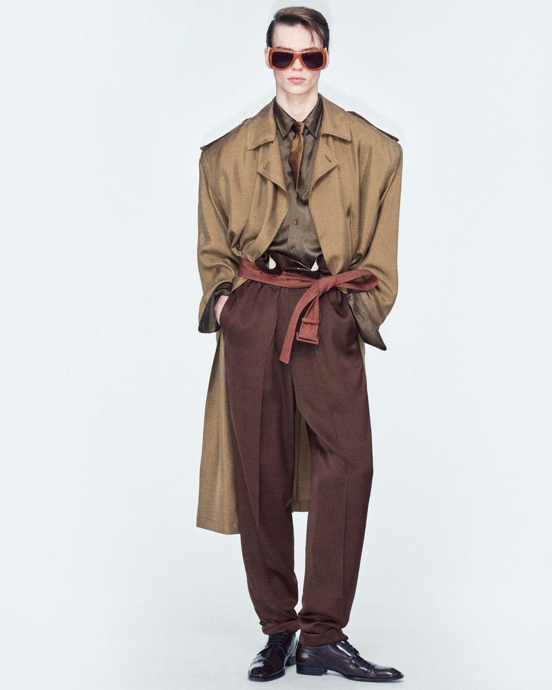 A singular adult male fashion model is indoors, wearing a brown coat, a brown shirt with tie, brown pants, a belt, sunglasses, and dress shoes.