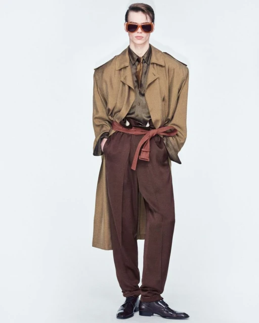 A singular adult male fashion model is indoors, wearing a brown coat, a brown shirt with tie, brown pants, a belt, sunglasses, and dress shoes.
