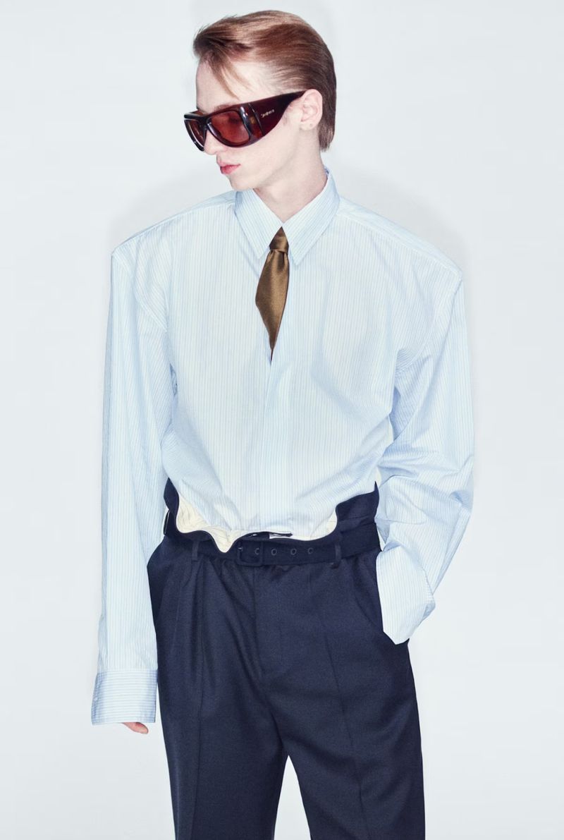 Model wearing dark wrap sunglasses, an oversized light blue striped shirt with broad shoulders, a tie, and dark trousers.
