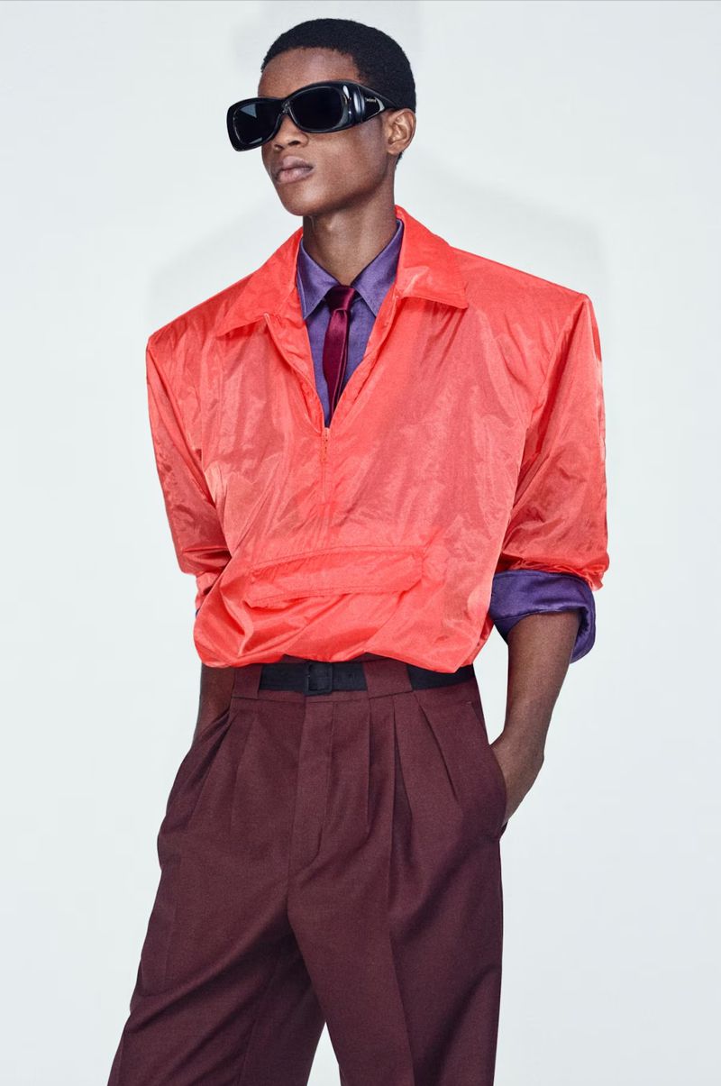 Young man in a coral shirt and maroon pants, posing against a white backdrop, accessorized with black sunglasses for a fashion-forward portrait.