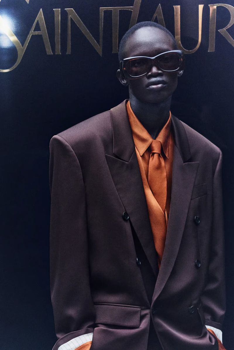 Fashion shot of a Black male model in a brown suit and orange shirt, wearing sunglasses, posing in front of the Saint Laurent logo.