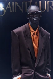 Black man wearing oversized brown blazer and orange shirt with sunglasses against dark background