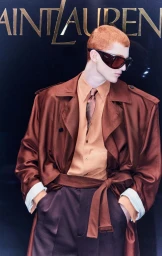Model with red hair wearing sunglasses, brown coat, and tie against a dark wall with Saint Laurent text