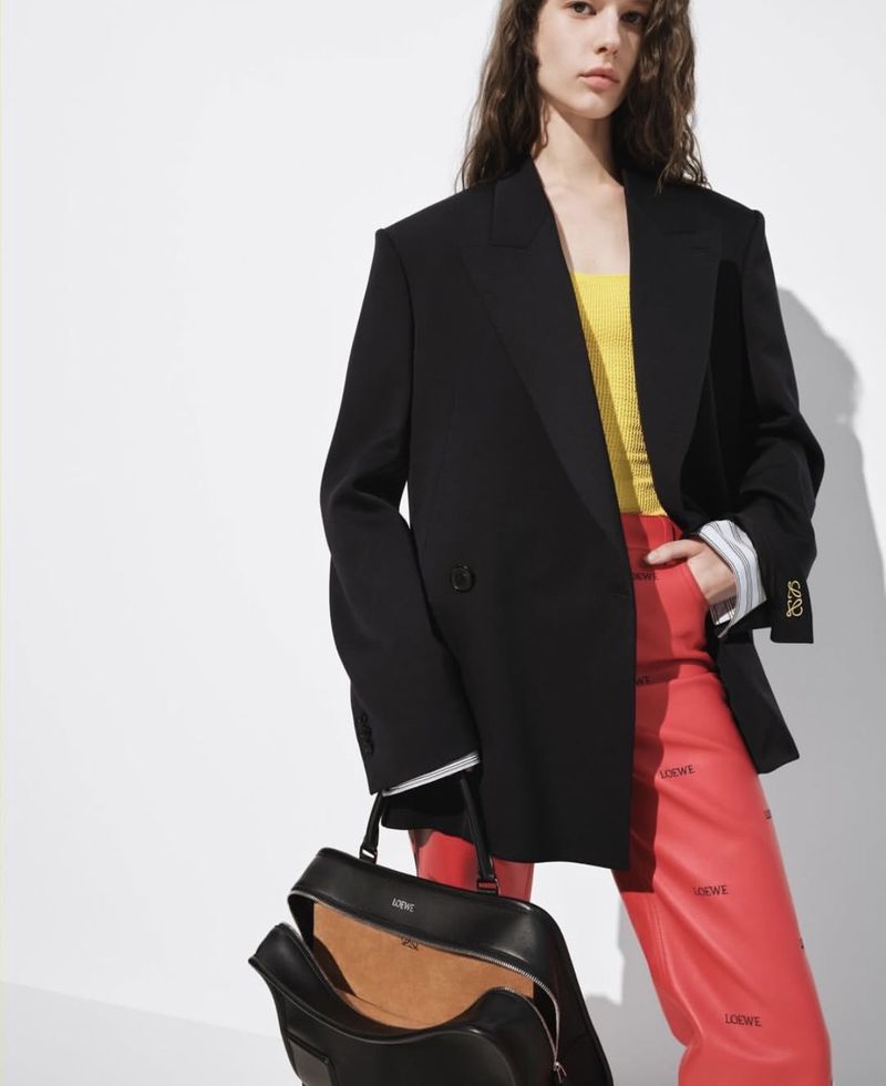 Fashionable woman in a black blazer, yellow top, and red printed pants, posing with a black Loewe handbag against a white background.