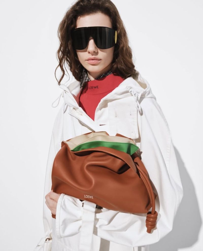 A woman wearing sunglasses holds a Loewe pouch bag in front of a white background. The model is wearing a white jacket and red top.