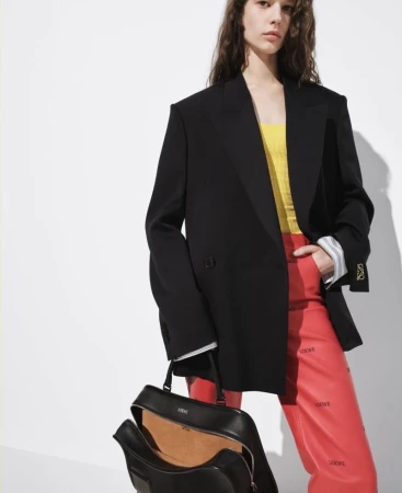 Model wearing an oversized black blazer, yellow knit top, and red printed trousers, holding an open black leather Loewe handbag.
