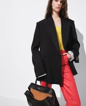 Model wearing an oversized black blazer, yellow knit top, and red printed trousers, holding an open black leather Loewe handbag.
