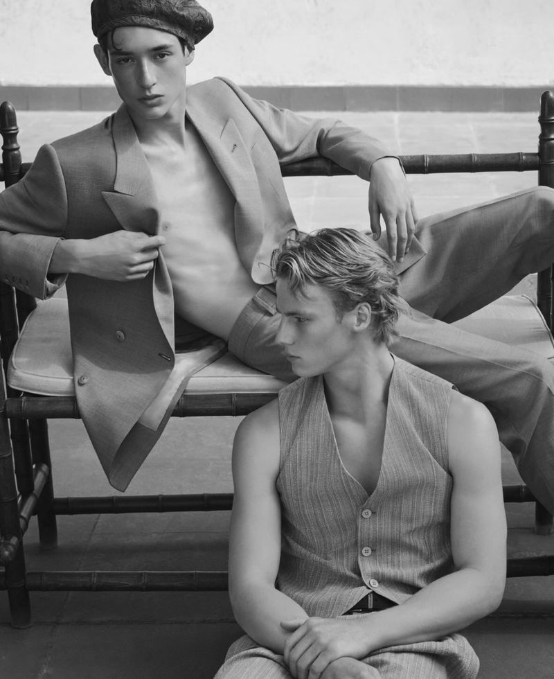 Two adult male models in matching tailored suits posed in a monochromatic indoor fashion editorial shot. One sits while the other reclines.