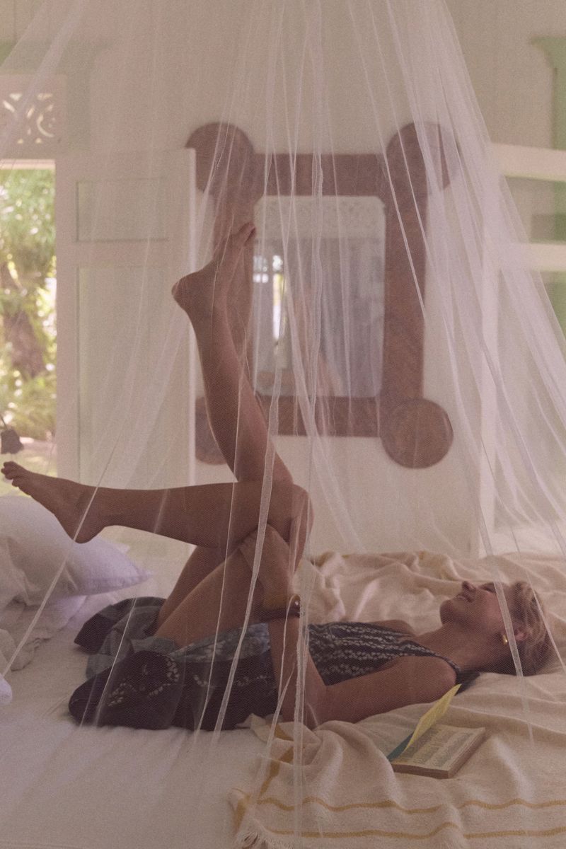 Woman relaxing on a bed with white mosquito netting in a brightly lit bedroom with a book, light sheets, and outdoor views.