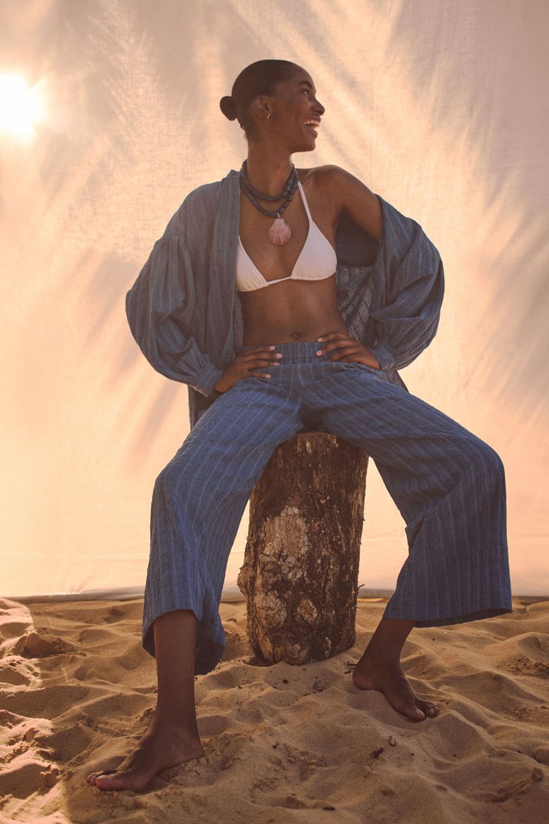 Woman in blue outfit posing on a beach log seat. She is wearing a white bikini top with layered bead necklaces in bright sunlight.