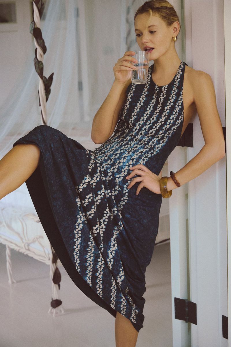 Young woman in a halter dress poses in an indoor setting while holding a glass of water and leaning against a white door frame.