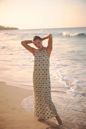 Model in a patterned green and cream maxi dress posing barefoot on a sandy beach with ocean surf in the background.