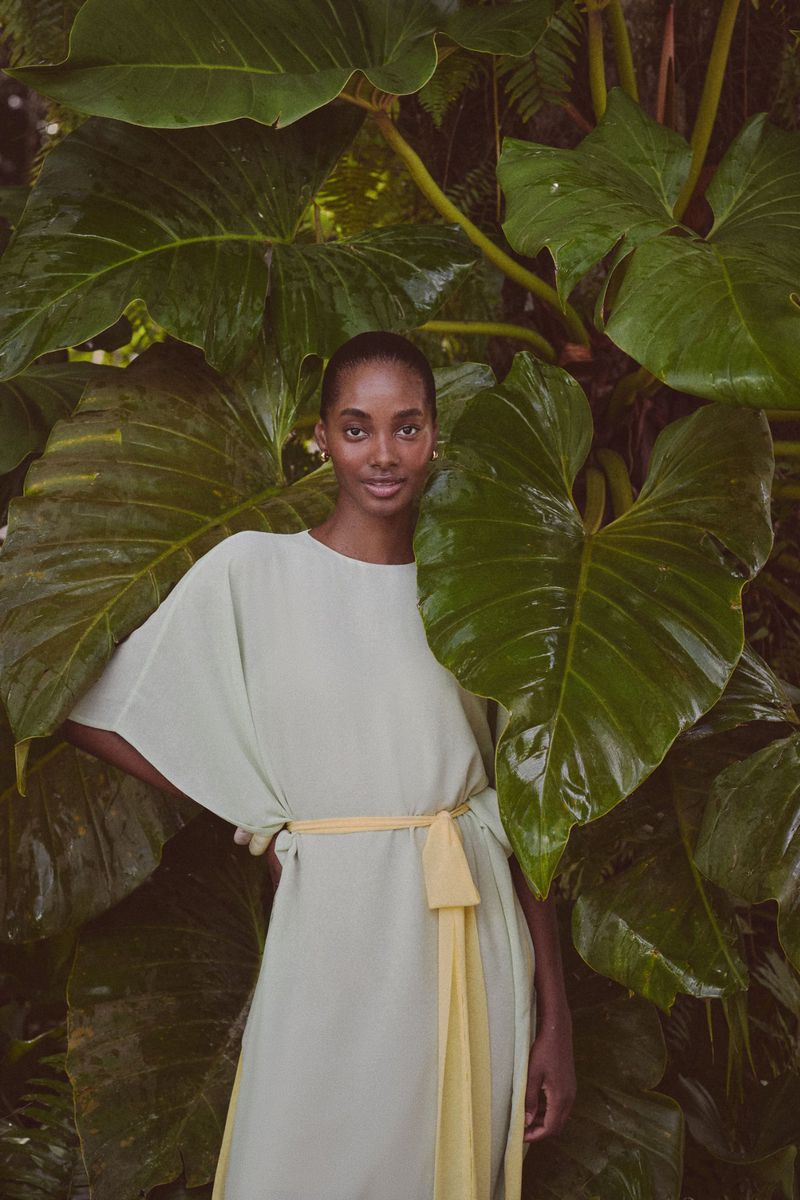 Elegant Black woman in flowing mint-green dress with yellow sash posing outdoors with tropical philodendron leaves in the background.