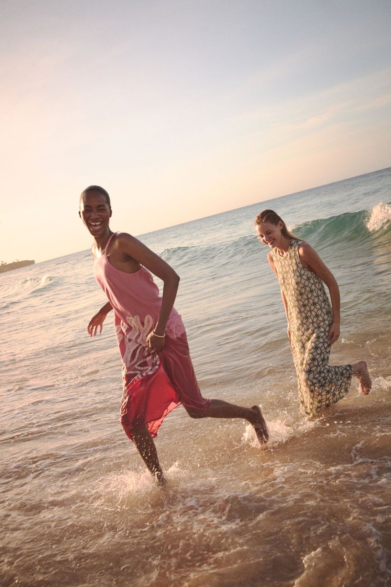 Two women in dresses laughing and running in ocean surf on a sunny day. One woman is African American and the other is Caucasian.