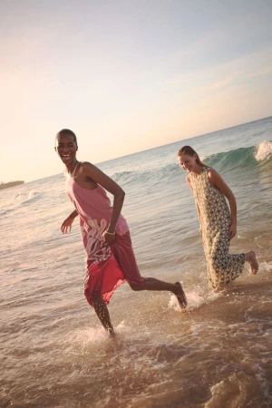 Two women in dresses laughing and running in ocean surf on a sunny day. One woman is African American and the other is Caucasian.