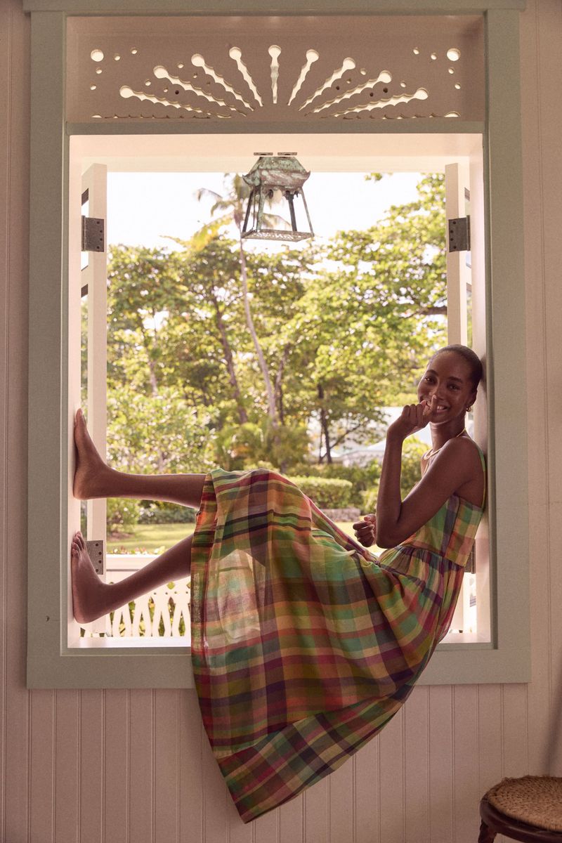 Woman in plaid dress sits in bright window with trees outside. Relaxed pose and bare feet add to the casual and summery aesthetic.
