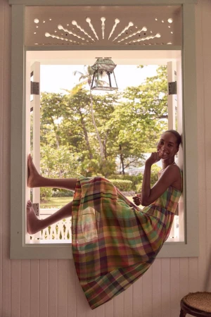 Woman in plaid dress sits in bright window with trees outside. Relaxed pose and bare feet add to the casual and summery aesthetic.